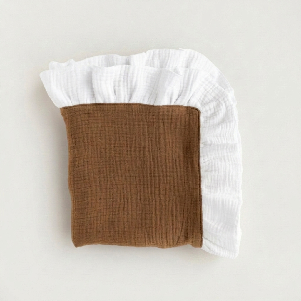 A folded muslin blanket with a brown center and white ruffled edges, photographed on a clean off-white background.