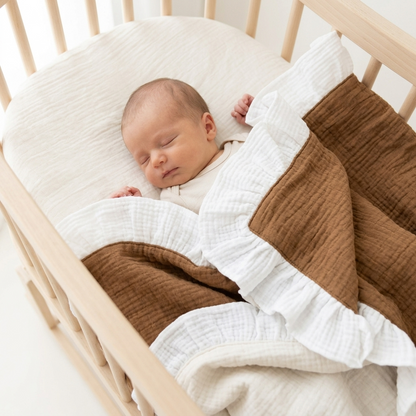 A sleeping newborn baby wrapped in a brown and white muslin blanket with ruffled edges, lying in a light wood crib.