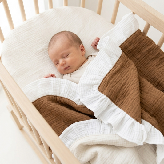 A sleeping newborn baby wrapped in a brown and white muslin blanket with ruffled edges, lying in a light wood crib.