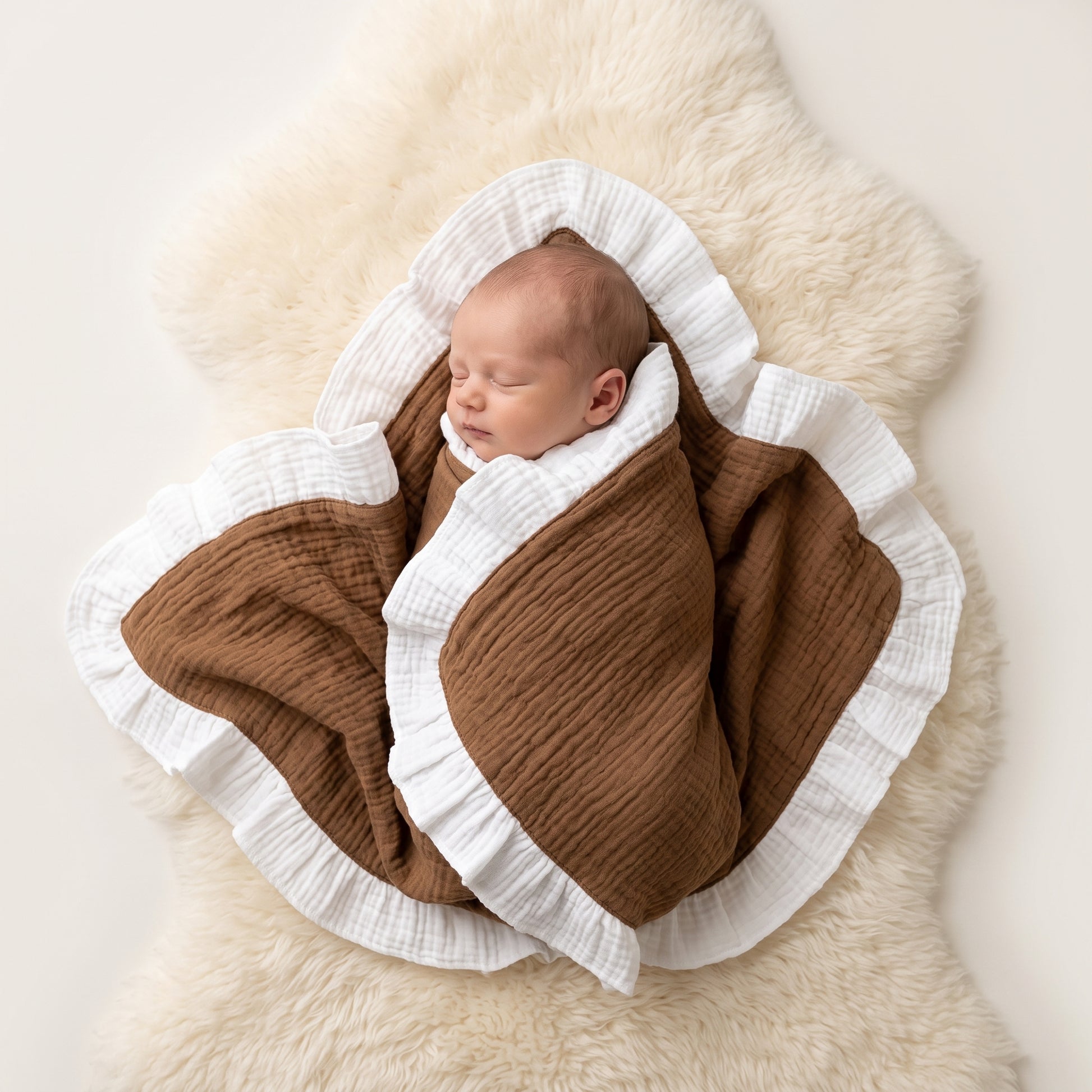 A sleeping newborn baby swaddled in a brown and white muslin blanket with ruffled edges, lying on a fluffy cream sheepskin rug against a light neutral background.