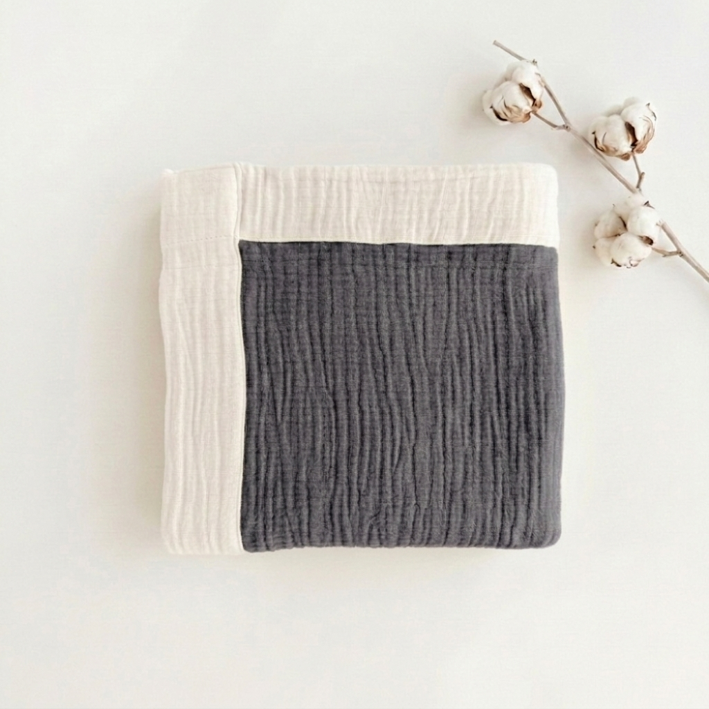 A folded textured muslin blanket in dark gray and off-white, styled with a cotton boll branch on a clean white background.