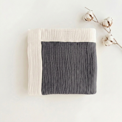 A folded textured muslin blanket in dark gray and off-white, styled with a cotton boll branch on a clean white background.