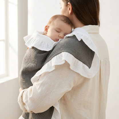 A woman wearing a light beige linen shirt carries a sleeping baby in a dark gray textured muslin wrap with white ruffled trim, set against a bright, softly lit indoor background.