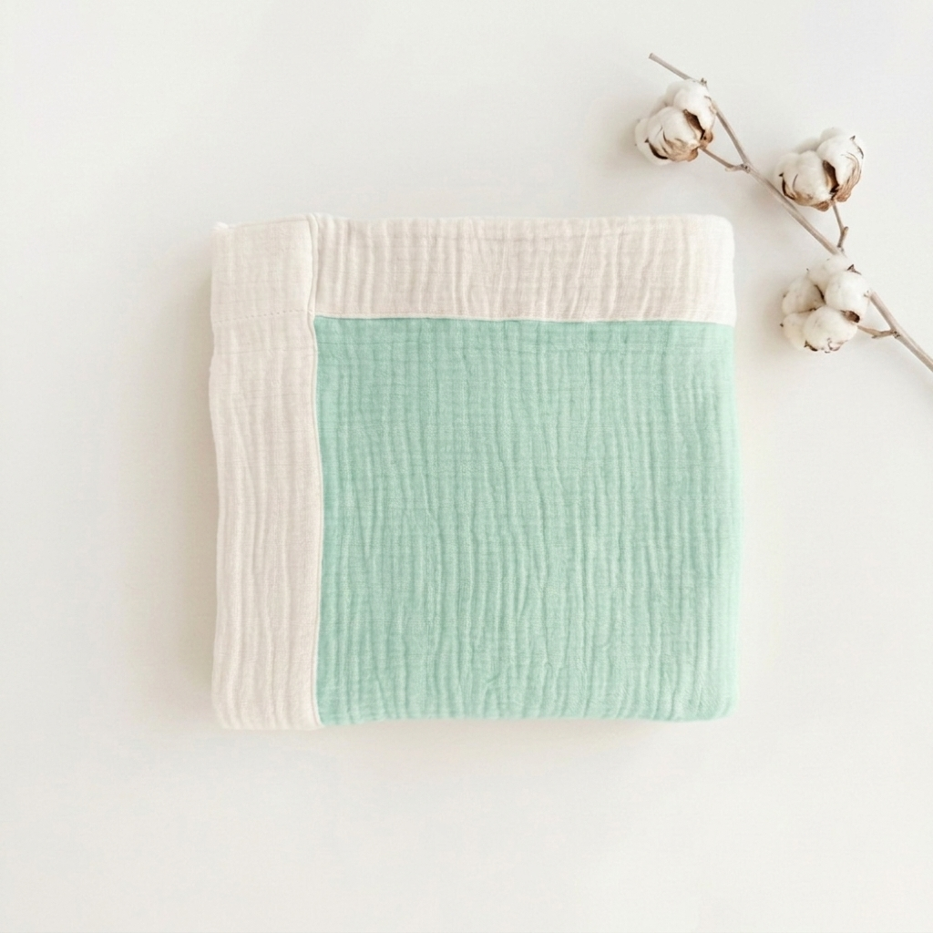 A folded muslin baby blanket with a mint green center and cream borders, styled on a white background with cotton bolls.