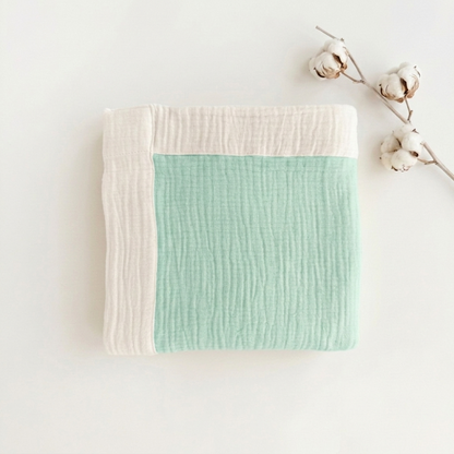 A folded muslin baby blanket with a mint green center and cream borders, styled on a white background with cotton bolls.