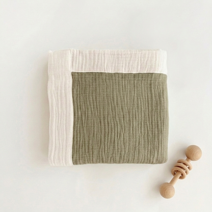 A folded muslin baby blanket in sage green and off-white, styled flat-lay with a wooden teething toy on a clean white background.