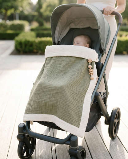 An olive green muslin baby blanket with white trim and wooden beads is draped over the footmuff of a stroller, with a baby resting inside and a mother's hand visible pushing the stroller on a wooden deck.