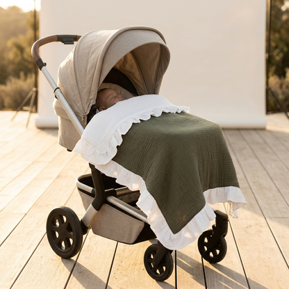 A sleeping baby is tucked inside a stroller outdoors on a wooden deck. The stroller is covered with a sage green textured blanket and a white ruffled muslin throw, with a beige canopy and stroller liner.