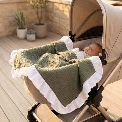 A baby sleeping in a beige stroller, covered with an olive green and white ruffled muslin blanket on a wooden deck.