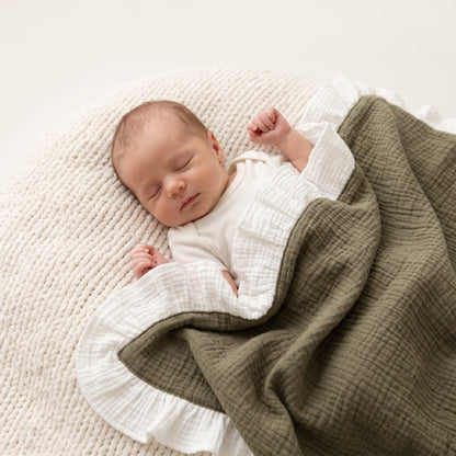 A sleeping newborn baby wrapped in a white long-sleeve bodysuit and an olive green muslin blanket with white ruffled trim, lying on a textured cream knit blanket.