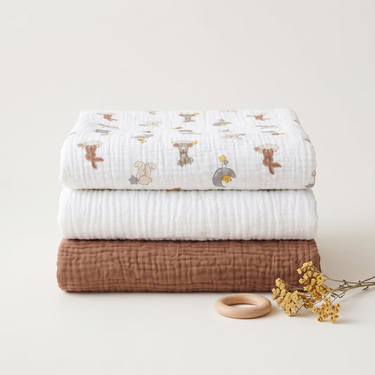 Three folded muslin baby blankets stacked on a light beige background, with a small wooden ring and dried yellow flowers beside them. The top blanket is white with a soft cartoon animal print in brown, beige, and gray, the middle blanket is solid white, and the bottom blanket is solid warm brown.