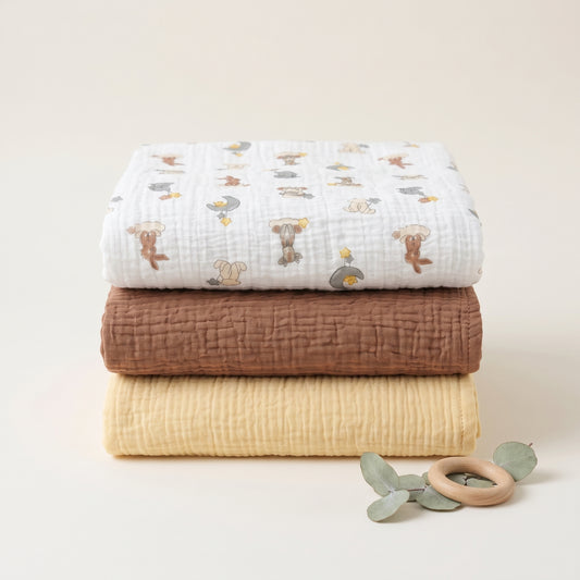 Three folded muslin blankets in white, brown, and pale yellow are stacked on a light beige background, with a small wooden teething ring and green leaves in front. The top blanket has a playful koala and bird pattern.