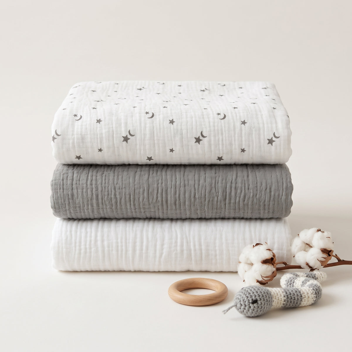 Three folded muslin blankets stacked vertically on a light beige background, featuring a white blanket with gray crescent moons and stars, a solid medium gray blanket, and a solid white blanket. A wooden ring toy and a crochet snake toy with cotton bolls are styled in front.