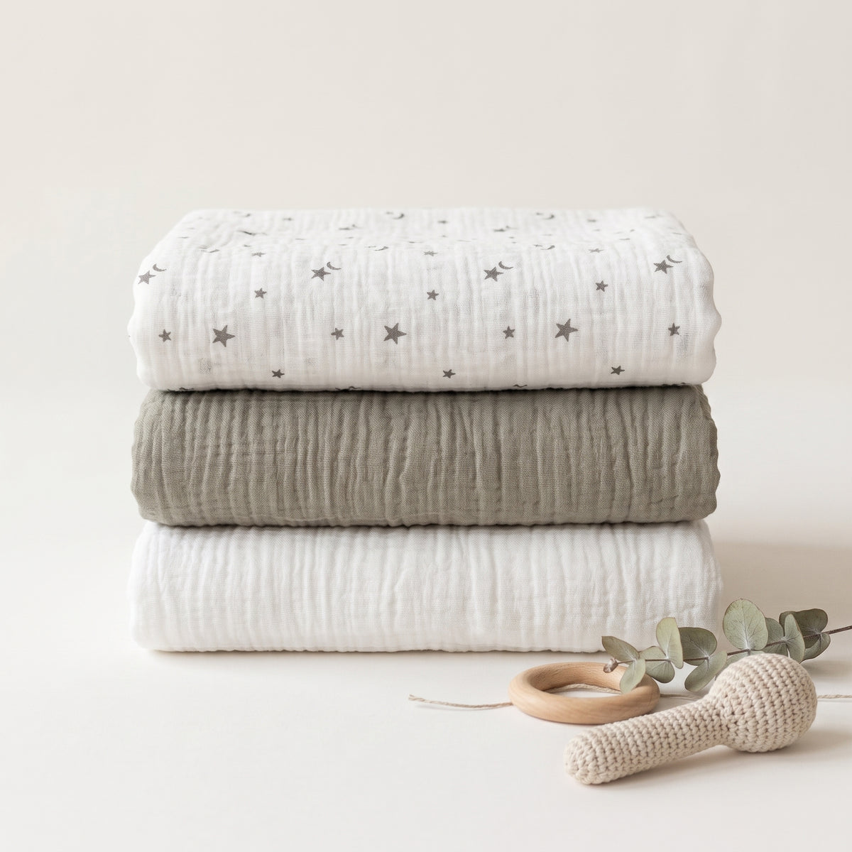 Three folded muslin blankets stacked vertically on a light beige background, with a wooden teething ring, crochet rattle, and eucalyptus leaves in the foreground. The top blanket is white with small gray star prints, the middle is a solid light gray, and the bottom is white.