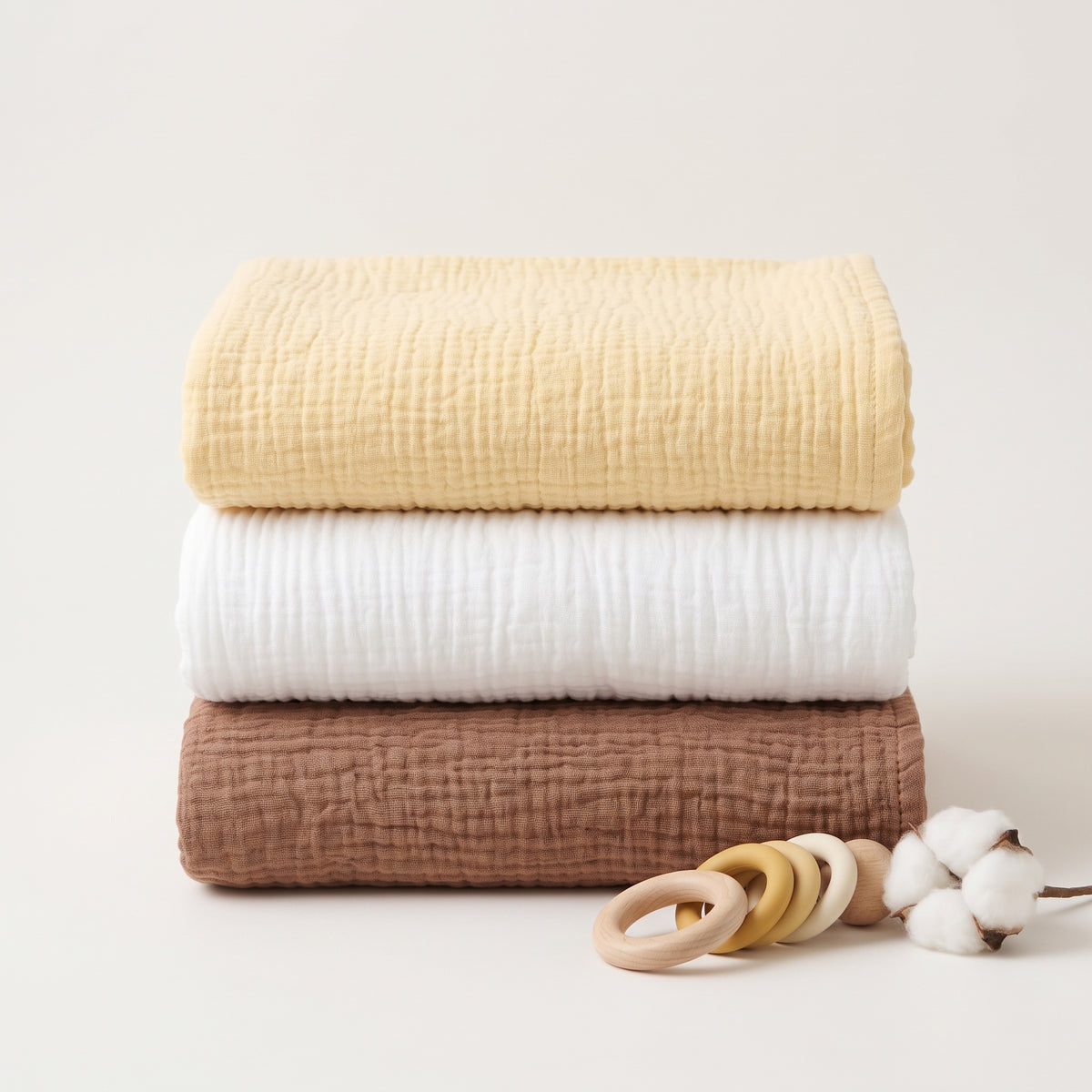 Three folded textured muslin blankets in pale yellow, white, and warm brown are stacked on a light neutral background, with a wooden ring teether and cotton bolls as styling props.