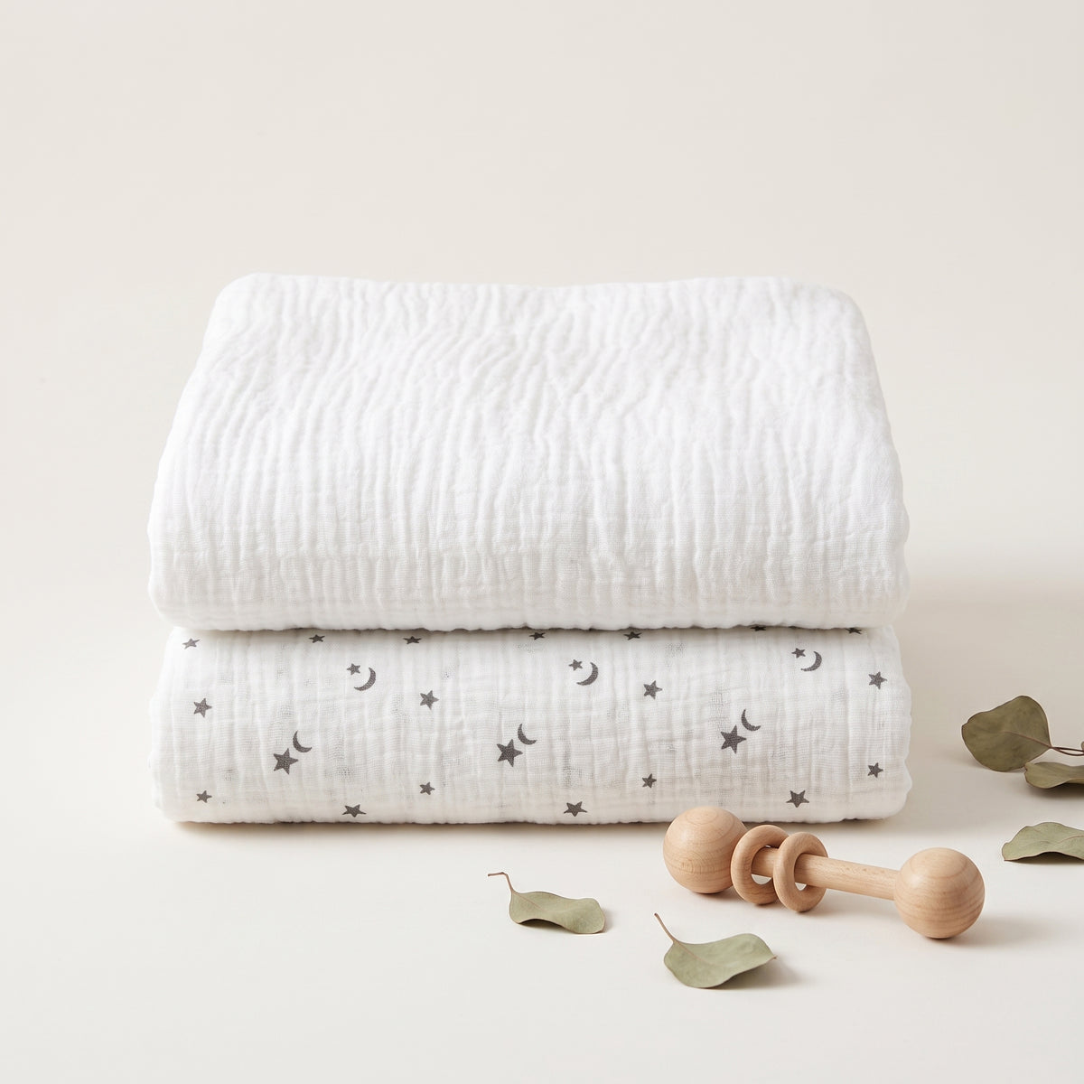 Two folded white muslin baby blankets stacked on a light beige surface, one plain and one with a small gray crescent moon and star pattern. A wooden baby teether and scattered dried leaves are placed in front.