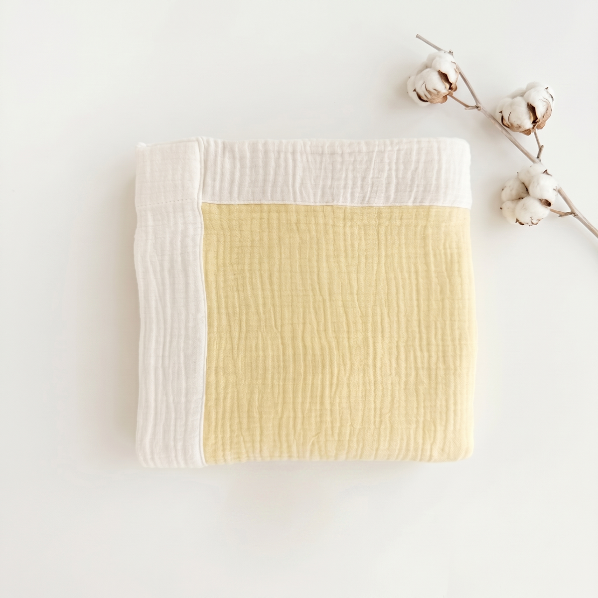 A folded muslin blanket with a pale yellow center and white borders, styled on a white background with cotton bolls.