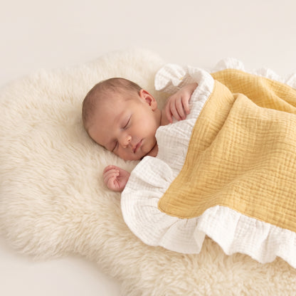 A sleeping newborn baby wrapped in a white ruffled muslin blanket with a mustard yellow side, resting on a fluffy cream sheepskin rug.