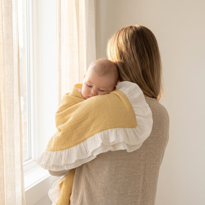 A woman from behind holds a sleeping baby wrapped in a soft muslin swaddle blanket. The blanket features a mustard yellow center with a white ruffled border, set against a bright, neutral room with a window and sheer curtains.
