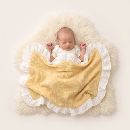 A sleeping newborn baby wrapped in a soft yellow and white ruffled muslin blanket, lying on a fluffy cream faux-fur rug against a warm off-white background.