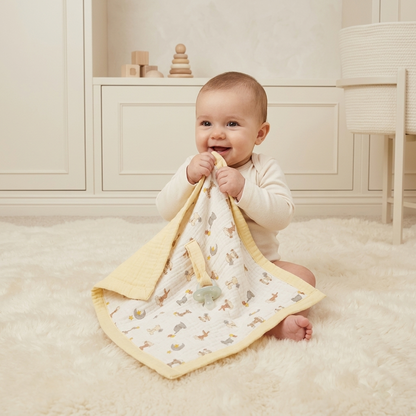 A baby sitting on a white fluffy rug holds a muslin security blanket with a yellow border and a small animal print. The baby is wearing a cream long-sleeve top in a bright, neutral nursery setting.