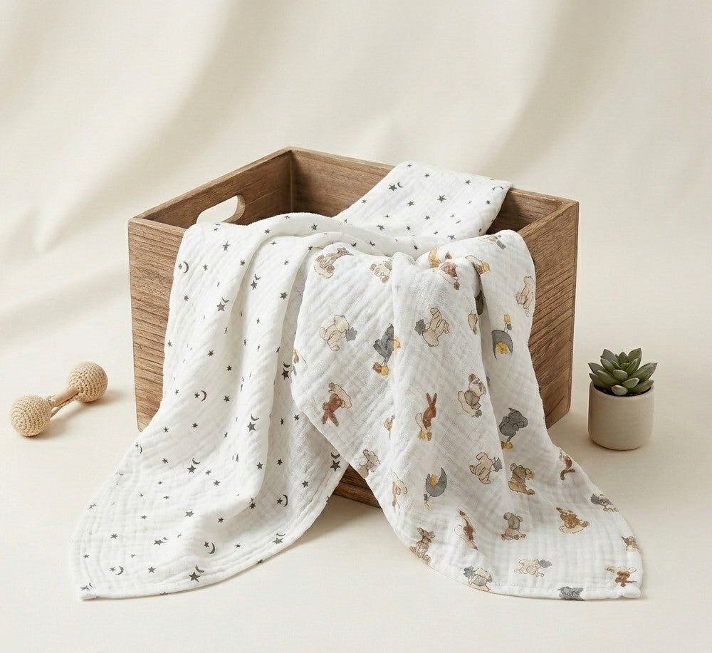 A white muslin baby blanket with a printed pattern  Dream Safari and Northern Stars  of bunnies, bears, and stars, draped over a wooden box with a matching folded piece behind it. The scene is styled with a wooden teething toy and a small potted succulent on a warm cream background.