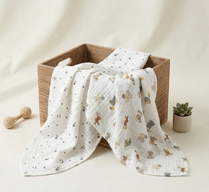 A white muslin baby blanket with a printed pattern  Dream Safari and Northern Stars  of bunnies, bears, and stars, draped over a wooden box with a matching folded piece behind it. The scene is styled with a wooden teething toy and a small potted succulent on a warm cream background.