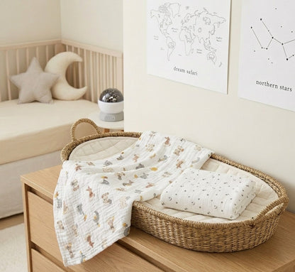 A woven oval baby bassinet basket on a wooden dresser, styled with a white patterned Dream Safari and Northern Stars muslin blanket and a folded white pillow with small gray dots in a softly lit nursery.
