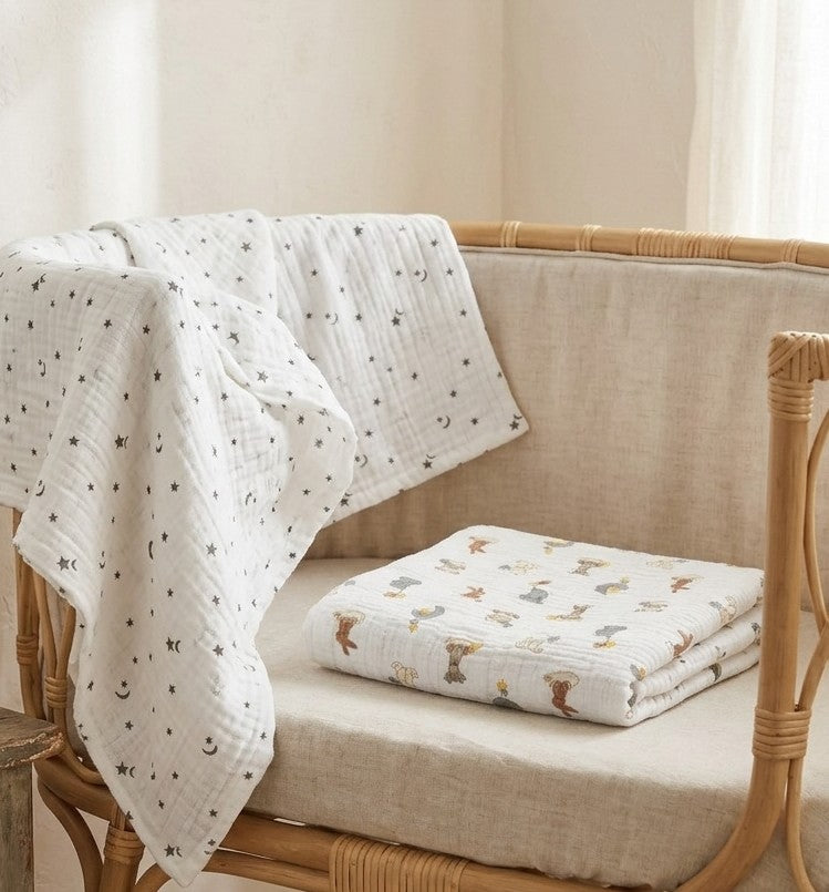 A white muslin blanket  Dream Safari and Northern Stars  with a black moon and star pattern draped over a woven rattan chair, with a folded matching blanket featuring a woodland animal print resting on the chair cushion.