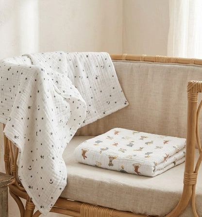 A white muslin blanket  Dream Safari and Northern Stars  with a black moon and star pattern draped over a woven rattan chair, with a folded matching blanket featuring a woodland animal print resting on the chair cushion.