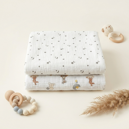 Two folded white muslin baby blankets with celestial and animal prints are arranged on a warm cream background with baby accessories including wooden teething rings, a plush toy, and dried pampas grass.