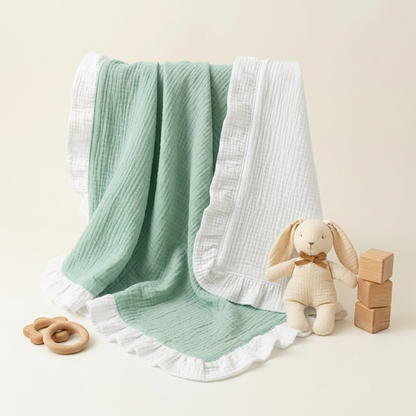 A mint  and white muslin baby blanket with ruffled edges, draped over a cream stuffed bunny toy next to wooden blocks and a wooden teething ring on a warm cream background.