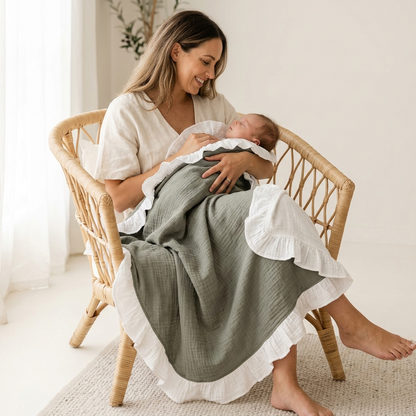 A woman sitting in a woven rattan chair holds a sleeping newborn baby wrapped in a olive grey muslin blanket with white ruffled trim, in a bright, neutral-toned room.